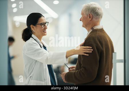 Médecin féminin caucasien saluant le patient masculin caucasien senior dans l'établissement médical, médecin souriant et plaçant la main sur l'épaule des patients, tout en tenant des lunettes dans l'autre main Banque D'Images