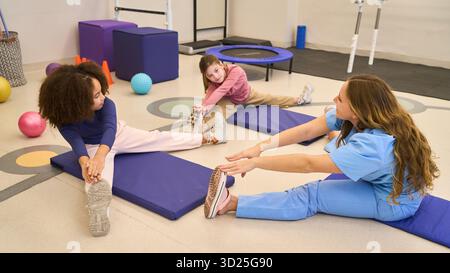 Un thérapeute guide les enfants à travers de doux étirements sur des tapis bleus dans un espace de rééducation pédiatrique, en utilisant des soins de soutien pour améliorer la motricité, l'équilibre, Banque D'Images
