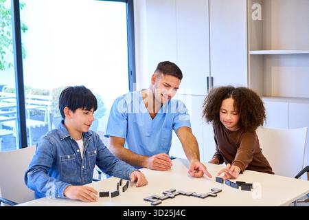 Un thérapeute en gommages bleus conduit deux enfants dans des activités motrices et d'équilibre à l'aide de blocs dans une clinique ensoleillée et accueillante. Soins de soutien ciblés Banque D'Images