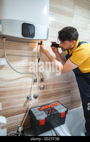 Plombier inspectant le chauffe-eau à l'aide d'une lampe de poche dans la salle de bains Banque D'Images