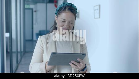 Debout mi adulte femme asiatique portant blazer beige tenant la tablette dans le couloir du bureau Banque D'Images