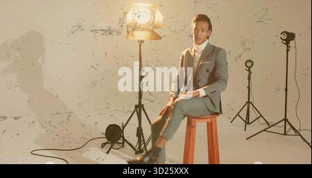 Homme asiatique portant un costume gris assis sur un tabouret dans le studio, avec des lumières, des cordes sur le mur peint-éclaboussé Banque D'Images