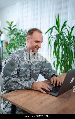 Homme militaire souriant travaillant sur ordinateur portable à la maison Banque D'Images