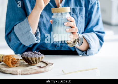 Femme boit Blue Iced Matcha Latte dans la cuisine près des accessoires pour sa préparation Banque D'Images