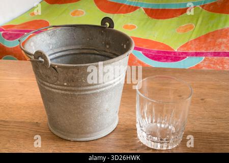 Verre vide et seau en métal galvanisé placé sur une surface en bois avec un fond coloré Banque D'Images