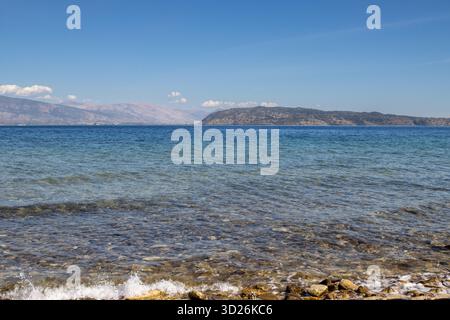 Eau propre et claire de la mer Ionienne, avec des rochers et des pierres visibles sur le fond. Montagnes albanaises à l'horizon. Ciel bleu avec des nuages blancs clairs. Banque D'Images