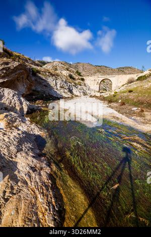 Une photographie couleur lumineuse, grand angle, capturant un lit de rivière sec avec un étroit jet d'eau qui coule sur des plantes aquatiques verdoyantes au premier plan Banque D'Images