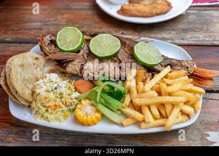 Un poisson entier frit garni de tranches de citron vert et accompagné de divers accompagnements sur une assiette blanche sur une table en bois. Banque D'Images