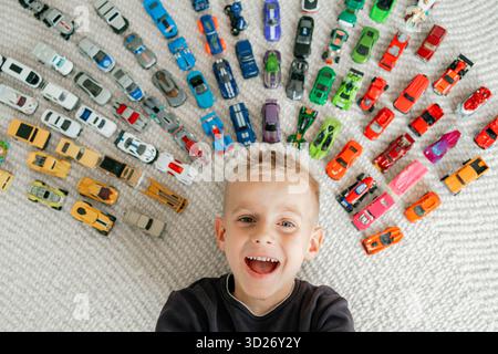 Photo descendante d'un garçon heureux souriant à l'intérieur d'un demi-cercle de voitures jouets colorées portrait d'enfance joyeux et collection de jeux Banque D'Images