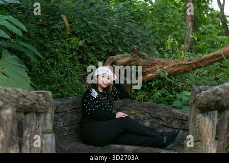 Un portrait d'une femme souriante dans un pull noir et béret blanc, se relaxant sur un banc de pierre entouré d'un feuillage vert luxuriant Banque D'Images