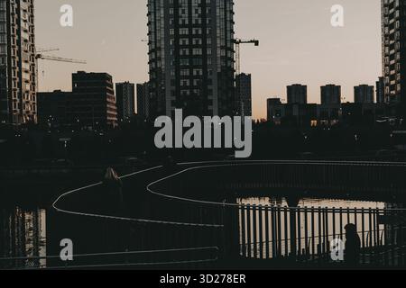 Paysage urbain en soirée avec des bâtiments modernes et un sentier sinueux près de l'eau. La silhouette d'une personne marchant le long d'un chemin est visible contre le b. Banque D'Images