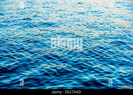 Gros plan de la surface de l'eau bleu calme avec des reflets doux de la lumière du soleil créant des ondulations dorées et des motifs de vagues abstraits Banque D'Images