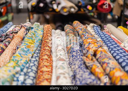 Gros plan de tissus enroulés soigneusement alignés dans un magasin, présentant des motifs floraux et abstraits vibrants dans les tons bleus, rouges, jaunes et blancs. Flou Banque D'Images