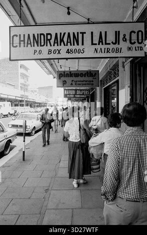 Nairobi, Kenya - Une scène de rue animée capture un moment de la vie quotidienne à Nairobi. Les clients font la queue devant Chandra Kant Lalji & Co, un établissement de premier plan dans le quartier commerçant de la ville. L'image montre une foule diversifiée de piétons, y compris des enfants, reflétant la population multiculturelle de la ville. La rue est pavée de briques, et les véhicules vintage ajoutent à l'ambiance historique. Cet instantané offre un aperçu du passé de Nairobi, mettant en évidence son activité commerciale dynamique. Banque D'Images