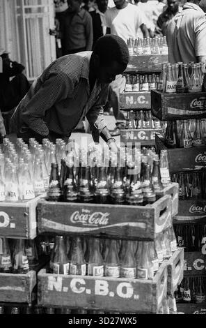 La vie de rue vibrante de Nairobi se déroule dans cette image en noir et blanc saisissante. Un travailleur solitaire trie méticuleusement les bouteilles de Coca-Cola empilées dans des caisses en bois, une scène emblématique du commerce animé de la ville. La photographie capture un moment d’activité quotidienne à Nairobi, au Kenya, mettant en valeur l’énergie brute du paysage urbain. Banque D'Images