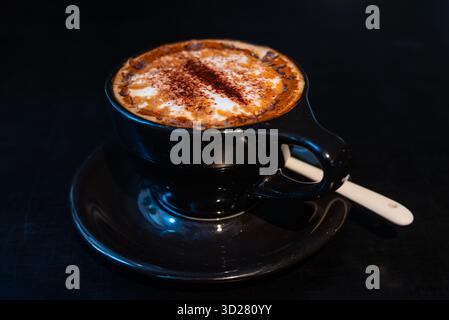 Une tasse de café fumant repose sur une table sombre. L'arôme riche remplit l'air tandis que la crème mousseuse et une pincée de cannelle créent un délice délicieux. Banque D'Images