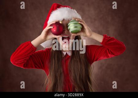 Fille souriante tenir la décoration de boules d'arbre de Noël dans ses yeux et porter le portrait de robe de chapeau de Père Noël en arrière-plan vintage Banque D'Images