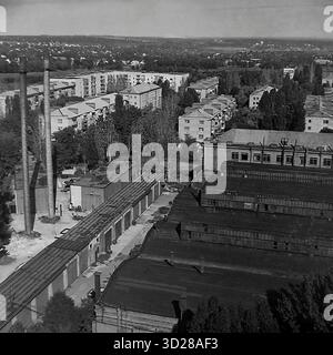 Archive photo noir et blanc de Sloviansk, Ukraine, août 1997. Cette vue en grand angle, prise 'du SKTB', montre un paysage urbain typique des années 1990 Le premier plan présente les toits d'une zone industrielle, une longue rangée de garages et deux hautes cheminées industrielles. En arrière-plan, séparé par des arbres, se trouve un micro-quartier résidentiel avec des bâtiments à panneaux de 5 et 9 étages. Cette image capture la proximité de l'industrie et de la vie résidentielle dans une ville post-soviétique. Banque D'Images