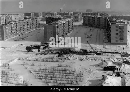 Archive photo en noir et blanc des années 1970 à Sloviansk, RSS d'Ukraine. Il s'agit d'une vue panoramique hivernale de la construction massive du microdistrict d'Artema. Tout le paysage est couvert de neige profonde. En arrière-plan, des dizaines de nouveaux bâtiments résidentiels à panneaux de 5 étages ('pyatietazhki') sont visibles. Le premier plan montre un chantier de construction actif avec des camions, des pièces de grue et des ouvriers. À gauche se trouve un jeune verger. Cette image documente le développement urbain soviétique à grande échelle. Banque D'Images