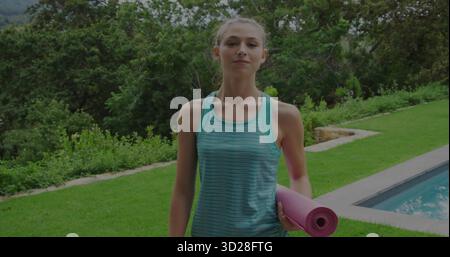 Femme debout portant des vêtements de sport, tenant un tapis de yoga rose sur la pelouse à côté du chemin de la piscine dans la cour ensoleillée Banque D'Images