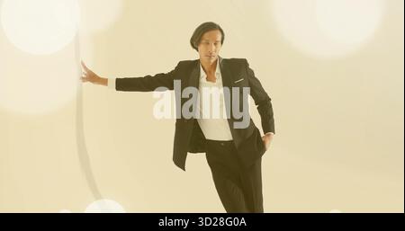 Modèle asiatique penché portant un blazer noir et un pantalon prolongeant le bras gauche en studio, lumières bokeh Banque D'Images