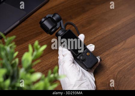 Poznan, Pologne - 16 octobre 2025 : DJI Osmo Pocket, un appareil photo compact à cardan noir, tenu par une main gantée, sur un bureau en bois pour le vlogging et la création Banque D'Images