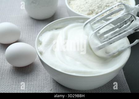 Blancs fouettés dans un bol, mixeur, farine et oeufs sur une table grise, gros plan Banque D'Images