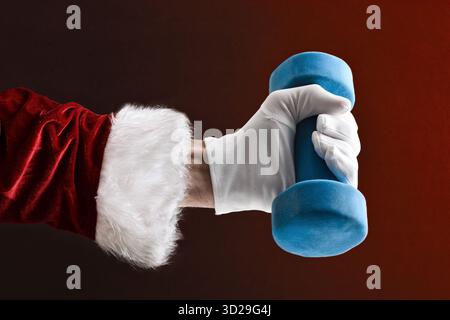 Main du Père Noël en costume et gants soulevant un haltère bleu sur fond isolé rouge. Banque D'Images