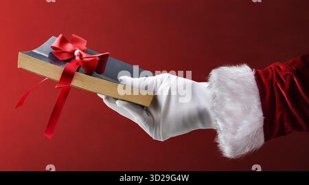 Concept de donner des livres de lecture avec la main du Père Noël portant des gants blancs tenant un livre de poche noir avec un arc rouge sur un dos rouge isolé Banque D'Images