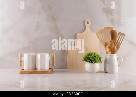 Deux élégantes tasses en céramique blanche, un set de planche à découper, un pichet avec des outils en bois et une fleur en pot sur un comptoir en marbre. fond de cuisine Banque D'Images