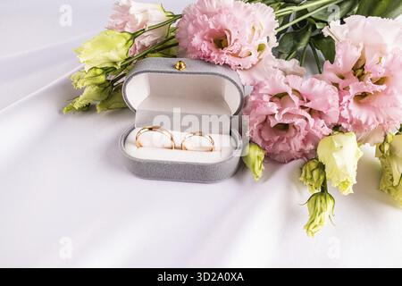 Deux anneaux classiques de mariage en or dans une boîte en velours gris sur un tissu de satin blanc avec des fleurs roses fraîches. Concept de mariage. Vue de face Banque D'Images