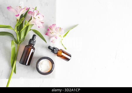 Deux flacons cosmétiques avec compte-gouttes avec produit naturel de soin du visage et un pot ouvert de crème sur un podium en marbre avec fleurs printanières. Vue de dessus Banque D'Images