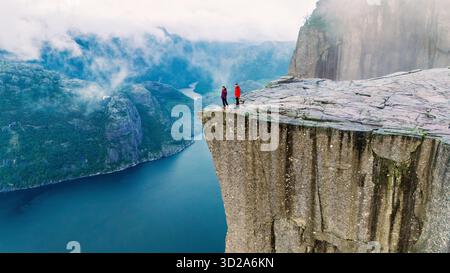 Deux randonneurs se tiennent au bord de la falaise Preikestolen, surplombant un magnifique fjord en Norvège. La brume matinale recouvre doucement le paysage, créant une atmosphère sereine et majestueuse. || autorisation du modèle Banque D'Images