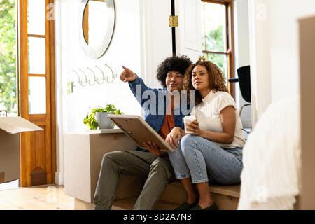 Jeune couple diversifié planifiant un décor à la maison tout en étant assis sur des boîtes mobiles, se sentant excité. Déménagement, déballage, design d'intérieur, style de vie, carton, famille || autorisation du modèle Banque D'Images