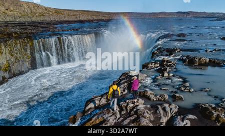 Deux aventuriers se tiennent sur un terrain rocheux près de Dettifoss, observant la majestueuse cascade Selfoss entourée d'un arc-en-ciel. Les couleurs vibrantes contrastent magnifiquement avec le paysage accidenté de l'Islande || modèle libéré Banque D'Images
