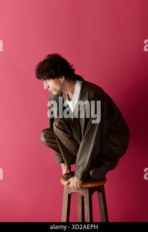 Un homme confiant dans une tenue élégante accroupie sur un tabouret en bois, exsudant charme et créativité. Banque D'Images