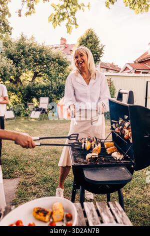 Femme âgée heureuse tenant la spatule debout près du barbecue dans la cour arrière Banque D'Images