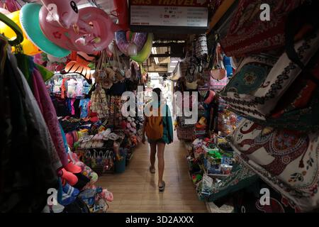 Femme en short avec un sac à dos marchant à travers un marché asiatique couvert avec des étals vendant du matériel de plage, des jouets et des vêtements Banque D'Images