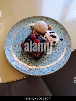 carré de brownie au chocolat garni de baies et de confiture accompagné de glace à la vanille et de crème fouettée, servi sur une assiette en céramique bleue. Banque D'Images
