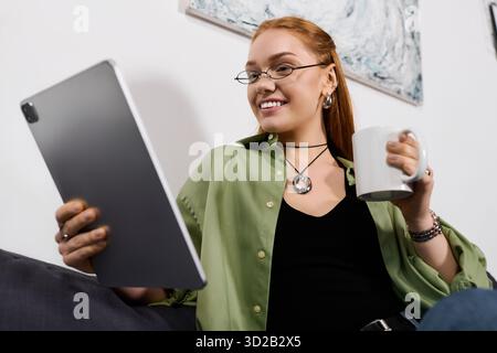 Une jeune femme joyeuse se détend à la maison, sirotant un café et utilisant sa tablette, entourée de confort. Banque D'Images