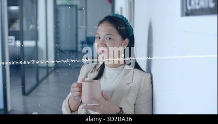 Femme asiatique adulte penchée en blazer beige tenant une tasse rose dans le couloir du bureau, avec forme d'onde audio Banque D'Images