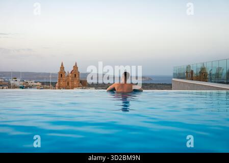 Une personne se repose près d'une piscine à débordement sur le toit à Mellieha, Malte au crépuscule, avec le Sanctuaire de notre-Dame de Mellieha et Comino et Gozo visible au-delà de CA Banque D'Images