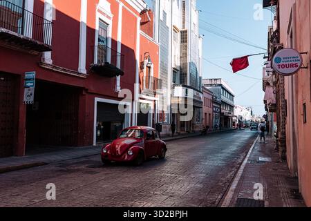 Merida, Yucatan, Mexique, Red Volkswagen Beetle est garé dans une rue de la ville. La rue est bordée de bâtiments et a quelques piétons à pied Banque D'Images