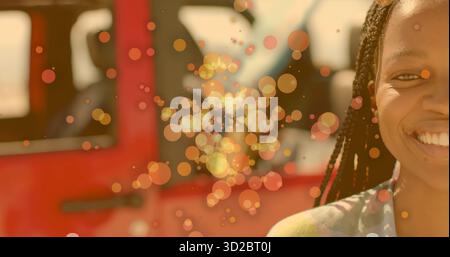 Femme avec des cheveux tressés souriant à côté de SUV rouge dans l'allée, avec bokeh orange-or, espace de copie Banque D'Images