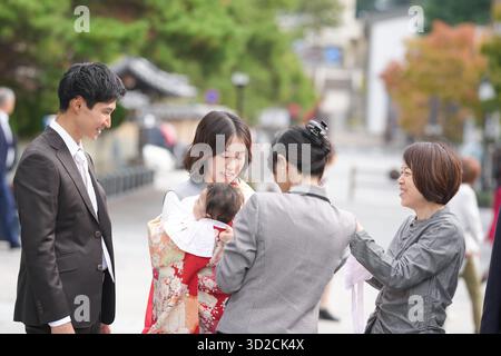 Un groupe de personnes se tient dehors, dont l'une tient un bébé. L'homme porte un costume et une cravate, et la femme porte une robe. La scène est c. Banque D'Images