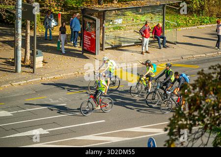 31 octobre 2025 , Belgrade, Serbie, cyclistes en gilets lumineux roulent le long d'une rue ensoleillée de Belgrade, en passant un arrêt de bus où plusieurs personnes se tiennent et si Banque D'Images