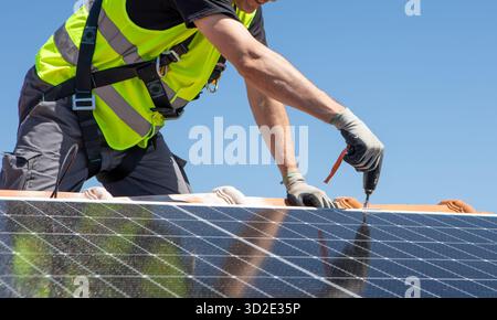Un travailleur portant un gilet de sécurité et des gants installe soigneusement un panneau solaire sur un toit sous un ciel bleu clair, mettant en valeur le changement en cours vers Renewa Banque D'Images