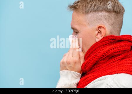 Un homme malade dans une écharpe et un chapeau se souffle le nez sur une serviette sur un fond bleu isolé. Le concept de rhume et de grippe dans la saison froide Banque D'Images