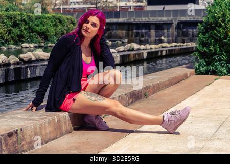 Femme avec des cheveux roses colorés et tatouage, portant des vêtements de sport, assise en toute confiance près d'un étang urbain dans un parc Banque D'Images