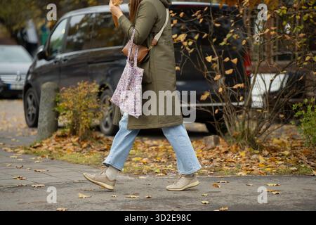 Personne marchant sur le trottoir de la ville en automne portant un manteau vert, un Jean clair et des baskets tenant un sac fourre-tout floral montrant le mouvement du style de vie urbain Banque D'Images